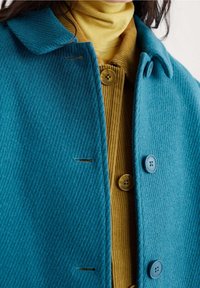 Grønblå frakke i uldblanding med en struktureret finish, der har krave, to blå knapper og en sennepsgul ribstrikket top med rundede knapper nedenunder.