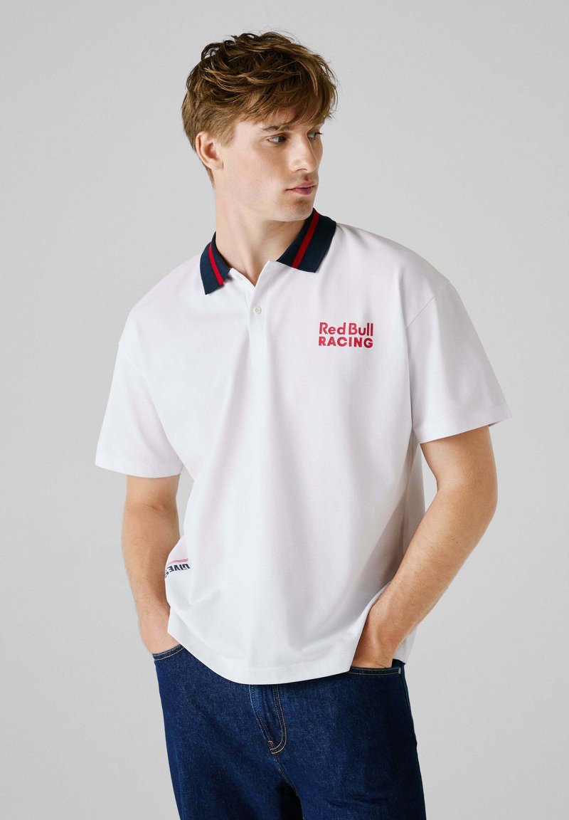 Hvid polo shirt med navy og rød stribet krave, med "Red Bull RACING" logo i rød. Korte ærmer og afslappet pasform, lavet af glat stof.