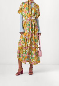 Blomstret kjole i gule, grønne og pink nuancer med korte ærmer, indsnævret talje med et hvidt bælte; parret med pink hæle og taske.