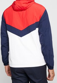 Red, navy, and white windbreaker jacket with a chevron pattern, hood, and elastic cuffs; lightweight, smooth texture; casual design.
