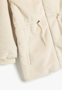 Veste en fausse fourrure de couleur crème clair, présentant une texture douce, des poignets côtelés et un cordon de serrage avec des finitions en métal doré. Comprend des poches latérales.