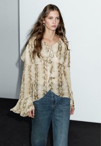 Beige ormönstrad blus med volangdetaljer, knytnacke och utsvängda ärmar, kombinerad med blå denimjeans. Minimalistisk bakgrund.