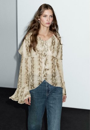 Blusa beige con stampa a serpente, dettagli smerlati, scollatura legata e maniche a campana, abbinata a jeans in denim blu. Sfondo minimalista.