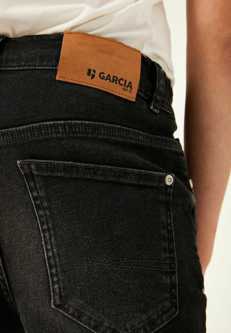 Jean en denim noir avec un patch en cuir marqué "GARCIA" sur la ceinture. Comprend des poches arrière et des accents de boutons en métal.