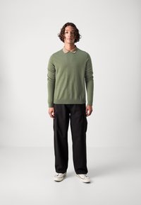 Světle zelený svetr s béžovým límcem, v kombinaci s černými cargo kalhotami. Materiál vypadá hladce a střih je volný. Obuv tvoří bílé tenisky.