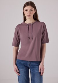 Mikina s krátkým rukávem, světle fialové barvy, s vysokým krkem, nastavitelnými šňůrkami a volným střihem, doplněná modrými džíny.