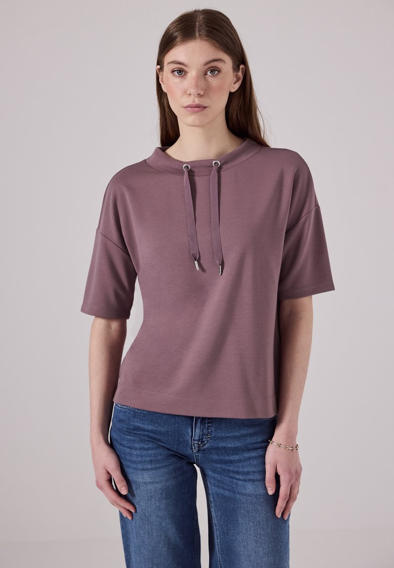 Mikina s krátkým rukávem, světle fialové barvy, s vysokým krkem, nastavitelnými šňůrkami a volným střihem, doplněná modrými džíny.