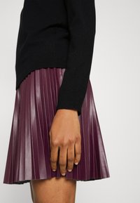 Femme portant un haut noir à manches longues et une jupe plissée en cuir violet, la main reposant doucement sur la jupe.