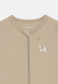 Bēšs kombinezons ar rāvējslēdzēju, apaļu kakla izgriezumu un mazu ziloni zīmējumu priekšpusē, kurš tur brūnu balonu. Mīksts kokvilnas materiāls.