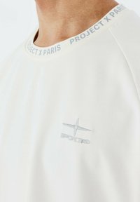 T-shirt a maniche corte di colore crema chiaro con scollo rotondo. Presenta un logo ricamato grigio sul petto e il testo "PROJECT X PARIS" sul colletto. Texture liscia.