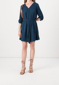 Robe sarcelle longueur genou avec un col en V, des manches trois-quarts et une ceinture assortie. Associée à des sandales à talons à brides couleur fauve.