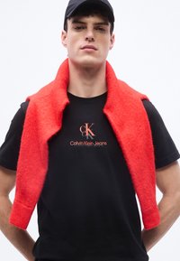 Νεαρός άνδρας με μαύρο μπλουζάκι Calvin Klein Jeans και μαύρο καπέλο, με ένα φωτεινό κόκκινο πουλόβερ περασμένο πάνω από τους ώμους του.