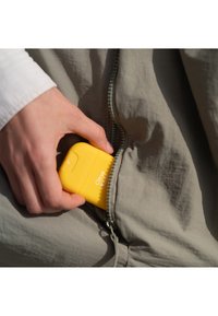Un oggetto a forma di agrume giallo che viene inserito in una tasca con zip di un indumento grigio chiaro. La tasca ha una zip verde.