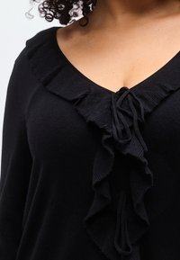 Pull noir à col en V profond, avec un volants froncés le long de l'encolure et un cordon de serrage à l'avant. Texture en maille douce.