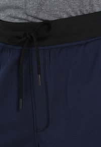 Närbild av marinblå byxor med svart elastiskt midjeband och dragsko knuten i en båge över grått tyg.