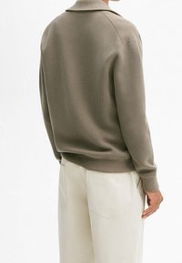 Bruine trui met kraag en geribde manchetten, gecombineerd met lichtgekleurde broek. Gladde textuur, aansluitende pasvorm en naadloze schouders.