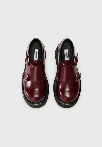 Ζευγάρι γυαλιστερών σκούρων κόκκινων δερμάτινων παπουτσιών με διπλούς γαντζοκαρφωτές, με παχιές μαύρες σόλες, που φαίνονται από ψηλά σε λευκό φόντο.