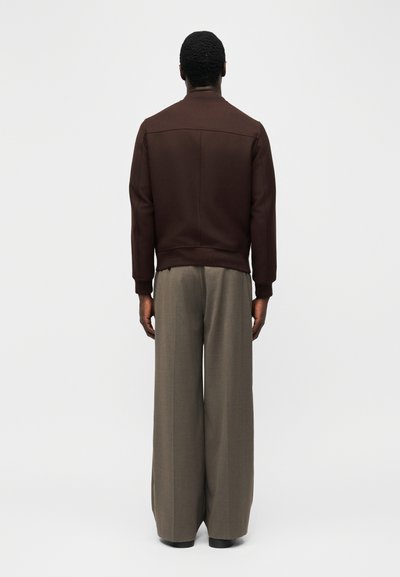 Άνδρας όρθιος με την πλάτη, φοράει σκούρο καφέ μπουφάν και ανοιχτόχρωμο γκρι παντελόνι με φαρδύ ραφή, σε απλό φωτεινό φόντο.