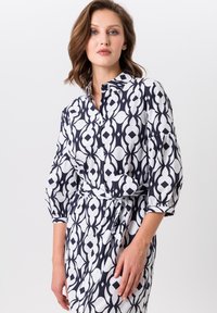 Robe boutonnée en bleu marine et blanc avec un motif géométrique, dotée de manches 3/4 et d'un détail de taille nouée pour une silhouette ajustée.