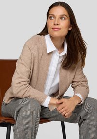 Hellbeige gestrickter Cardigan mit gerippten Kanten, getragen über einem weißen Hemd. Die Person sitzt und zeigt gemusterte graue und schwarze karierte Hosen.