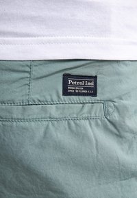 Pantalons en coton vert clair avec une poche arrière ornée d'une étiquette navy indiquant "Petrol Ind" et du texte sur le denim. Texture fine et lisse.
