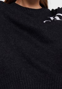 Svart stickad tröja med rund halsringning, ribbat slut och mjuk textur. Har vit logotypdetalj på framsidan. Nära bild.