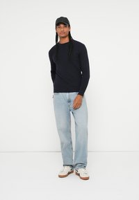 Pulover cu model canelat, navy, cu mâneci lungi, asociat cu blugi drepti de culoare albastru deschis și adidași albi cu talpă maro. Se poartă o pălărie neagră.