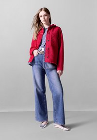 Röd texturerad jacka med svarta knappar, lager på lager över en jeansskjorta, ihop med vida blå jeans och silverfärgade balettklackar.