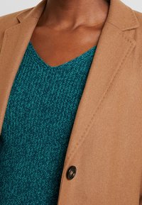 Brązowa marynarka na wymiar z wyciętym kołnierzem, zestawiona z teksturowanym turkusowym swetrem z dekoltem w serek. Mały czarny guzik jako akcent.