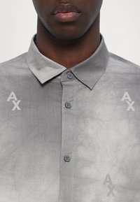 Homme portant une chemise grise boutonnée avec un col et de subtils logos "AX" sur la poitrine et les parties latérales inférieures.