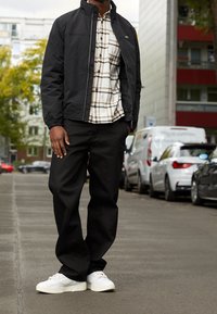 Man som står på en stadsgata klädd i svart jacka, rutiga skjorta, lösa svarta byxor och vita sneakers, med parkerade bilar i bakgrunden.