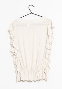 Blouse beige clair avec des manches froncées et une taille froncée. Présente un motif subtil et une texture douce. Accrochée à un crochet noir.
