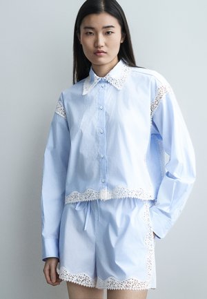 Νεαρή γυναίκα που φοράει ανοιχτόγαλάζιο πουκάμισο με κουμπιά και ταιριαστό σορτς με λευκή δαντέλα, στέκεται μπροστά σε μια απλή γκρίζα φόντο.