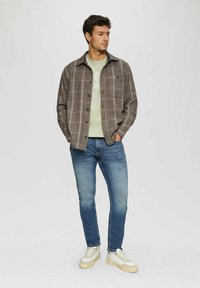 Gray plaid button-up shirt over a light green t-shirt, paired with blue jeans and cream-colored sneakers. Model stands in a neutral background.