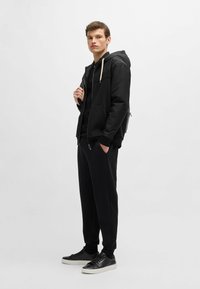Giacca nera con zip e cappuccio, abbinata a un top nero e pantaloni jogger. Completato con sneakers nere con suola bianca.