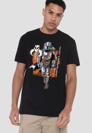 Junger Mann trägt ein schwarzes T-Shirt mit dem Mandalorianer in Rüstung, Grogu auf dem Rücken und weißem Mythosaur-Schädelsymbol auf orangem Hintergrund.