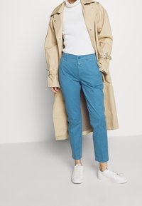 Beige trenchcoat över en vit polotröja, parat med turkosa byxor och vita sneakers. Byxorna är avsmalnande med en mjuk textur; knäppning med knapp.