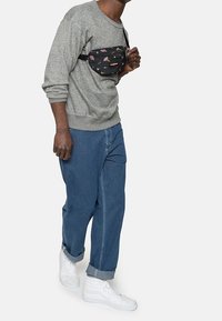 Svart midjeväska med blommönster, bärs över en grå sweatshirt och ljusblå denimbyxor, med vita sneakers.