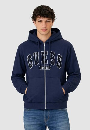 Giovane uomo con capelli ricci che indossa una felpa con cappuccio blu navy con zip e la scritta "GUESS 1981" e jeans blu, mani in tasca, sfondo neutro.
