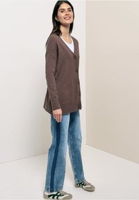 Cardigan marron avec col en V, fermeture à boutons, manches longues et coupe décontractée. Porté sur un haut blanc et associé à un jean bleu clair et des baskets.