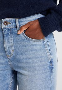 Světle modré džíny se slim fit střihem, obsahující kapsu, kontrastní stehy a hnědý knoflík v pase.