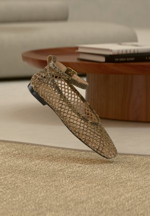 Chaussure plate avec un design en maille beige et une bride à la cheville, positionnée au-dessus d'un tapis tissé avec une table basse en bois et des livres empilés en arrière-plan.