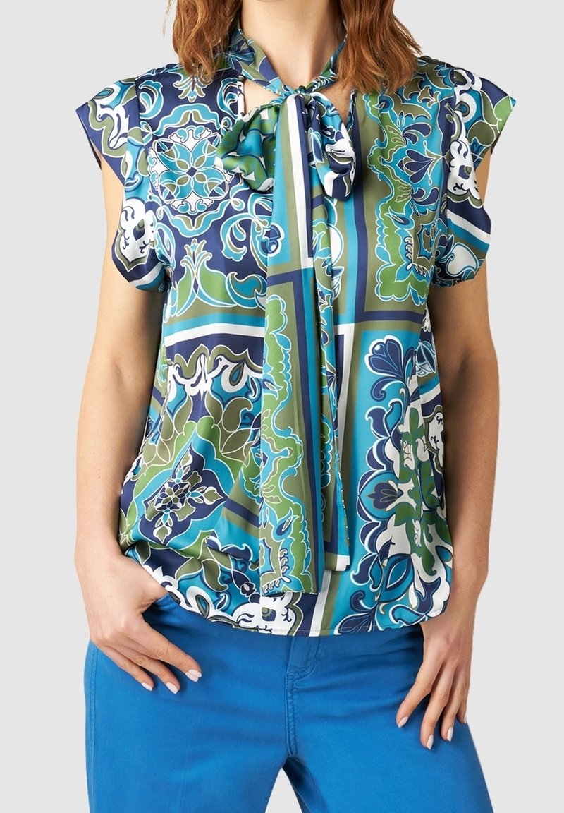 Femme portant un chemisier à manches courtes à motifs bleus, verts et blancs avec un foulard noué autour du cou et un pantalon bleu vif.
