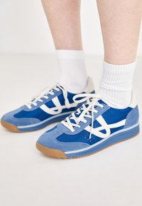 Zilā sporta apavi ar zamša un sietiņa augšdaļu, balti akcenti, plakani šņorēšanas vadi un gumijas zoles, kombinēti ar baltām rievojuma zeķēm.