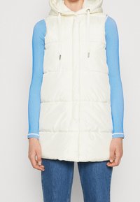White puffy vest with a hood, featuring drawstrings, quilted texture, and side pockets, layered over a blue long-sleeve ribbed top.
