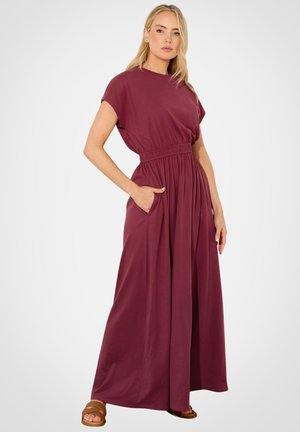 Vrouw draagt een mouwloze, hooggetailleerde, tot op de grond vallende bordeauxrode jurk met geplooide rok en zijzakken, staand tegen een effen achtergrond.