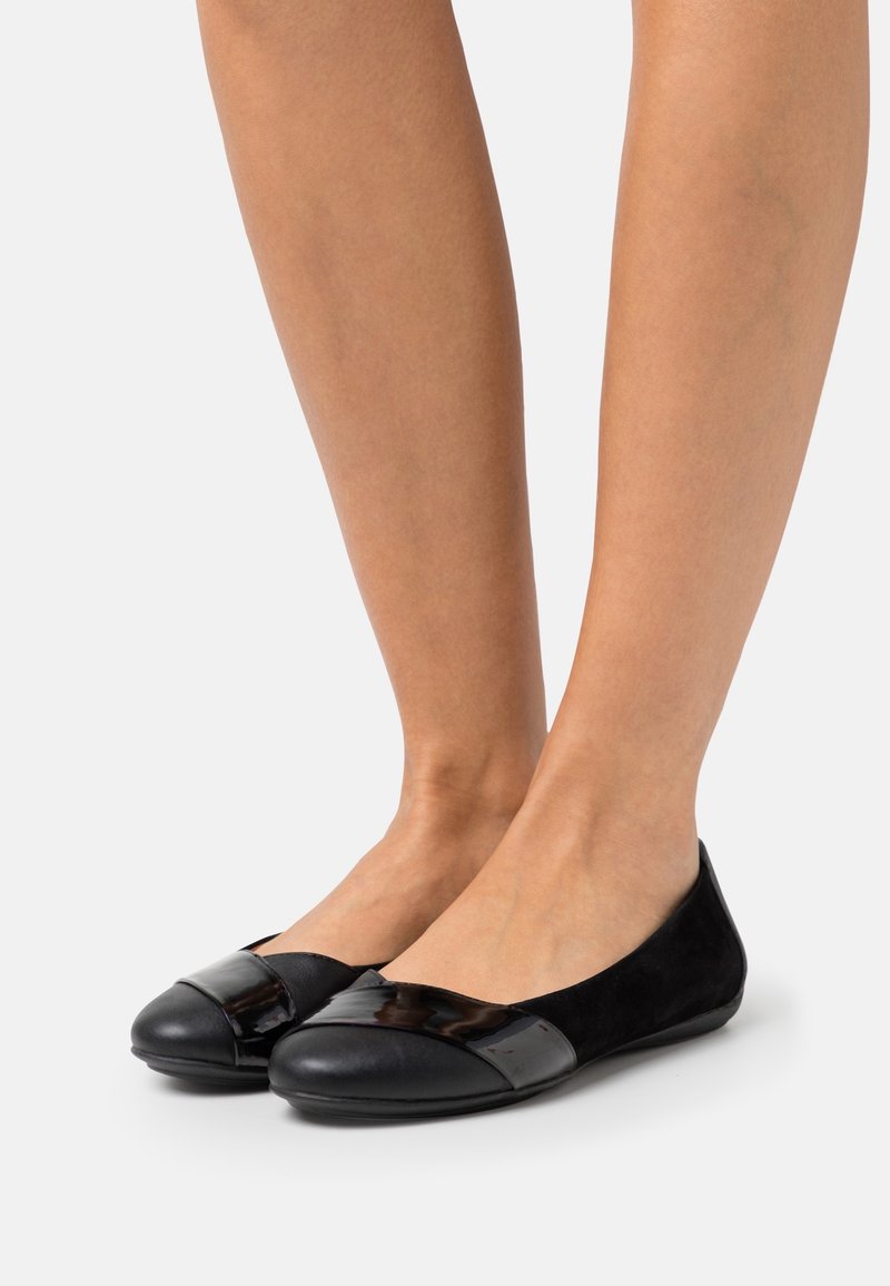 Jambes portant des ballerines noires avec un bout mat et un détail de bride brillante sur un fond clair uni.
