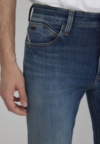 Jeans in denim blu scuro con vestibilità slim, dotati di una tasca frontale, un leggero sbiadimento e dettagli di cuciture arancioni. Classico design a cinque tasche.