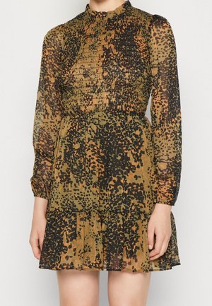 Kobieta w sukience z długim rękawem w nadruk misia, z marszczonym górnym stanem, falbankowym kołnierzem i warstwową spódnicą na jednolitym tle.