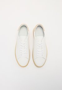 White leather sneakers with a smooth texture, featuring a beige suede heel tab, flat white laces, and a gum rubber sole.
